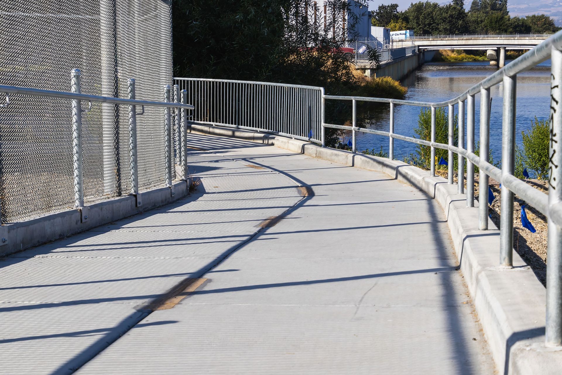 A curved pedestrian footbridge with metal railings along a river in an urban setting.