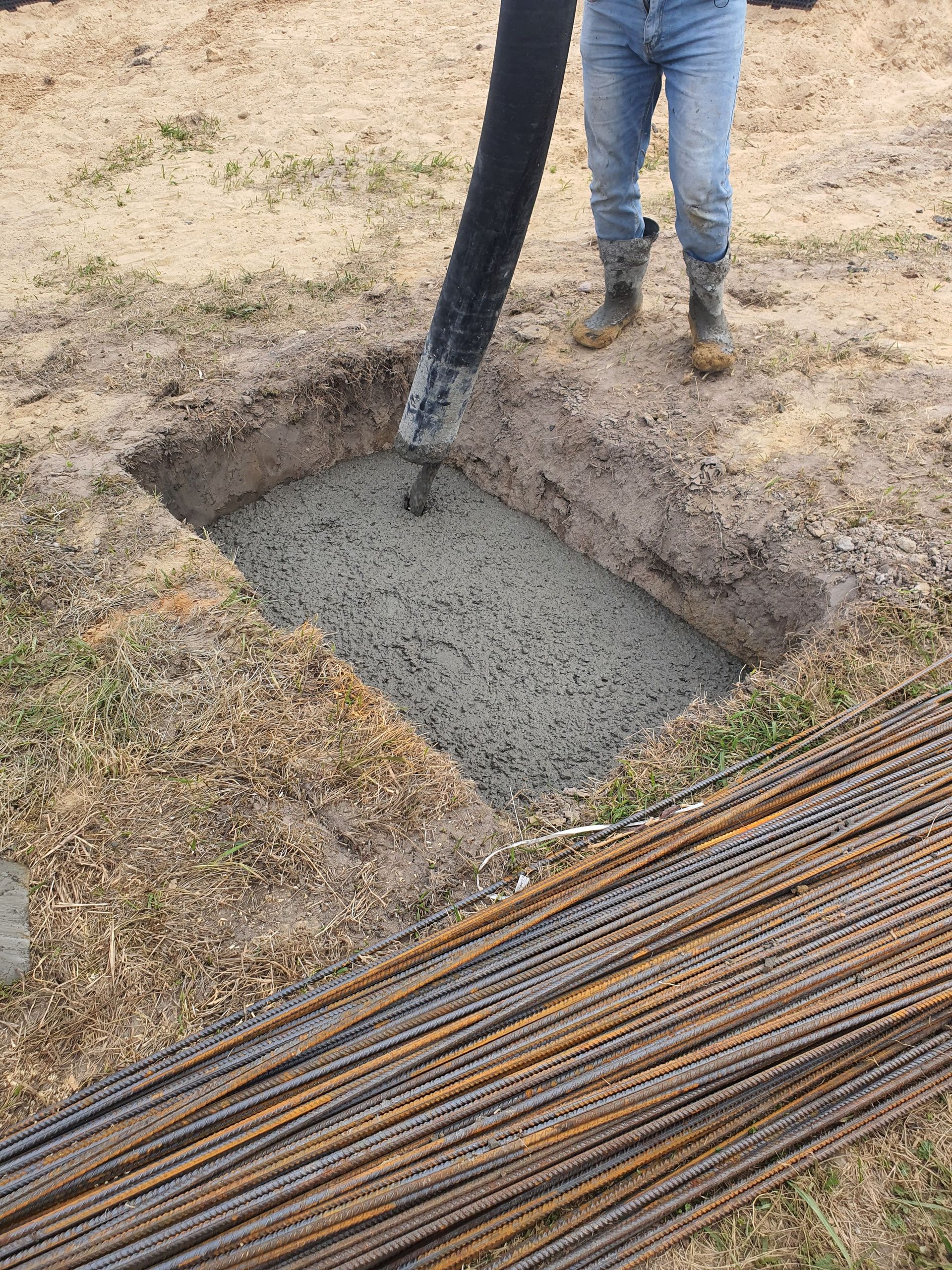 Concrete being poured into a foundation pit at a construction site in Poland, showing rebar and worker boots.